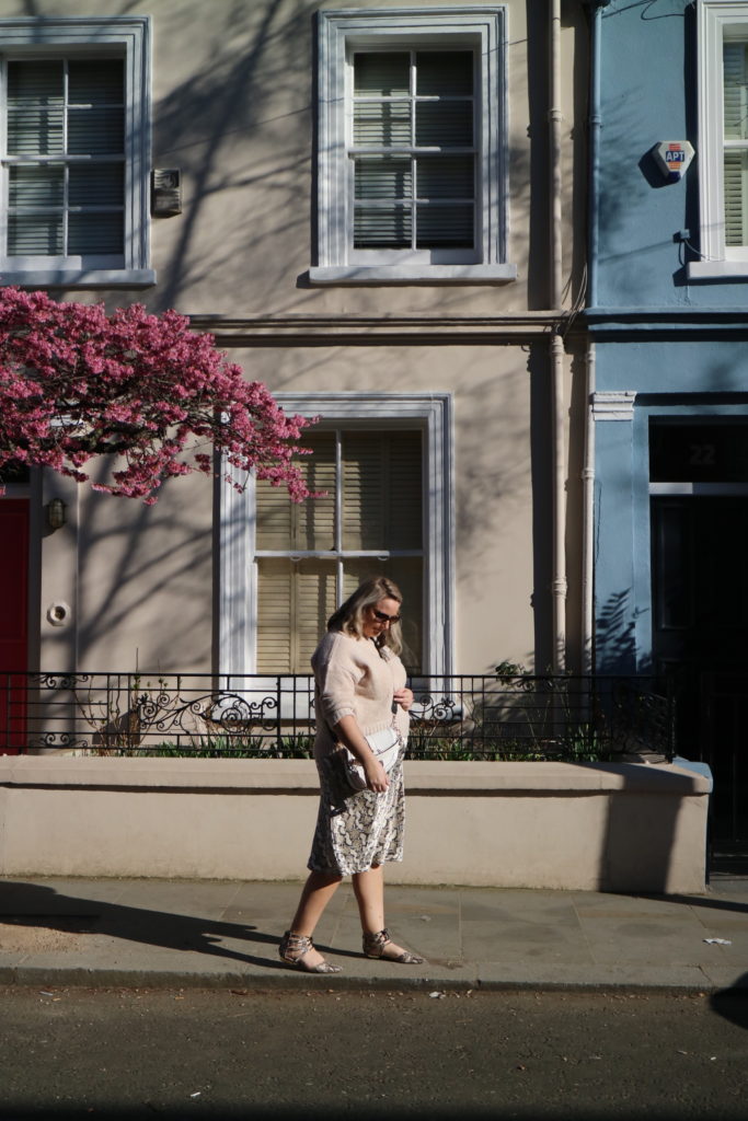 lucy walking down portobello road dressed in a satin snake print skirt, cream jumper and shoes and bag. in the background is a blossom tree and some coloured houses
